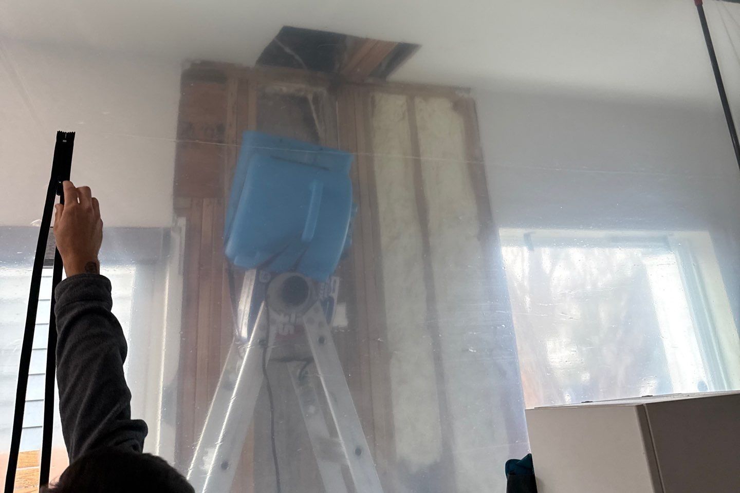 Plastic containment around an opened wall with exposed studs and insulation, and an air mover on a ladder drying the wall cavity in an Austin home